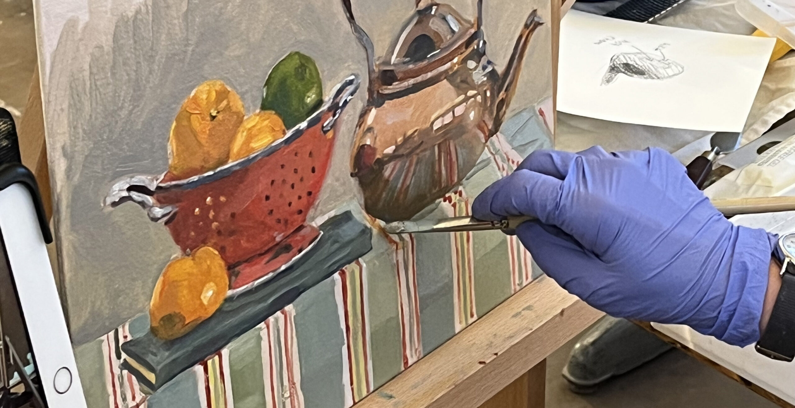 Blue-gloved hand holding paintbrush to still life oil painting in progress