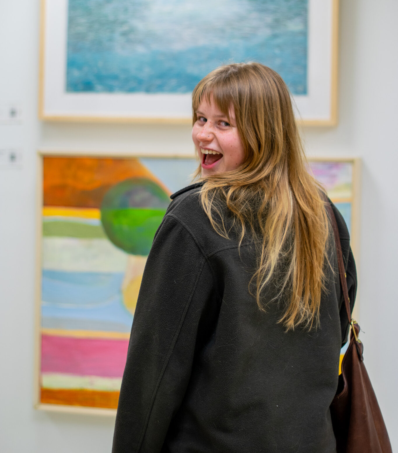 Long-haired person turning and grinning from looking at two artworks on the wall behind